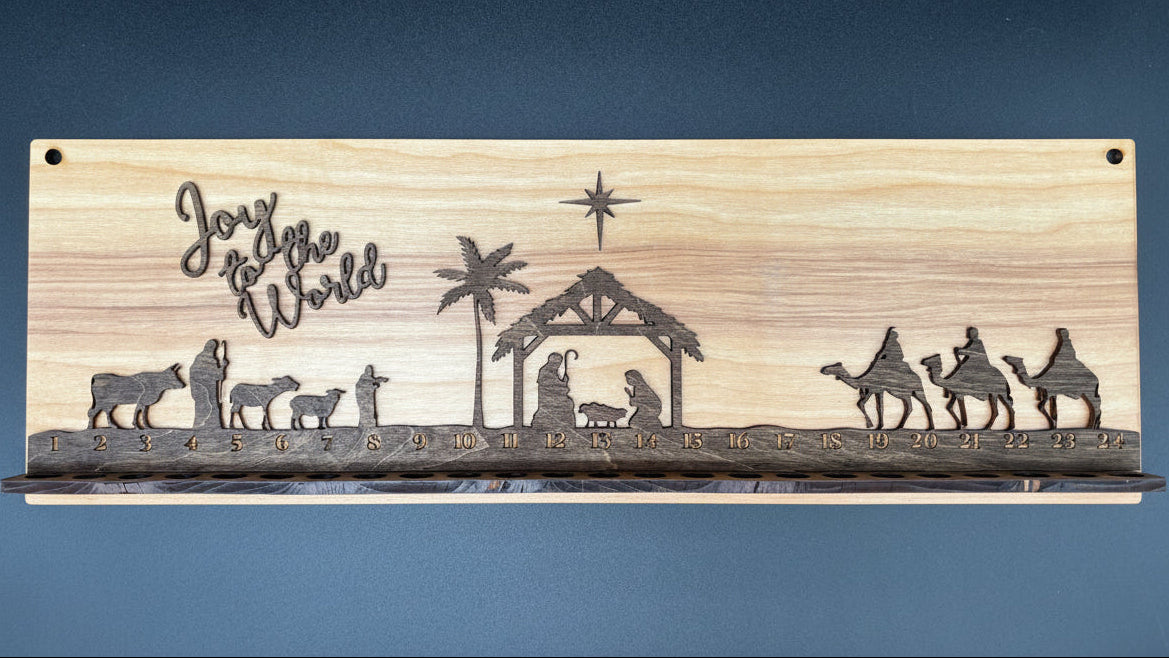 A rectangular wooden plaque featuring a laser-engraved Nativity scene with silhouettes of Mary, Joseph, baby Jesus, the stable, camels, and stars, alongside the text 'Joy to the World'. The plaque is shown against a dark slate-blue background.