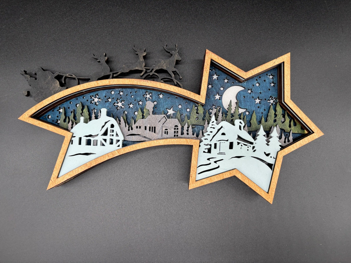 A star-shaped layered wooden cutout with a winter cabin scene, displayed on a dark gray background. The design features a light blue sky, white snow, green trees, and a dark blue night sky with stars and a crescent moon.