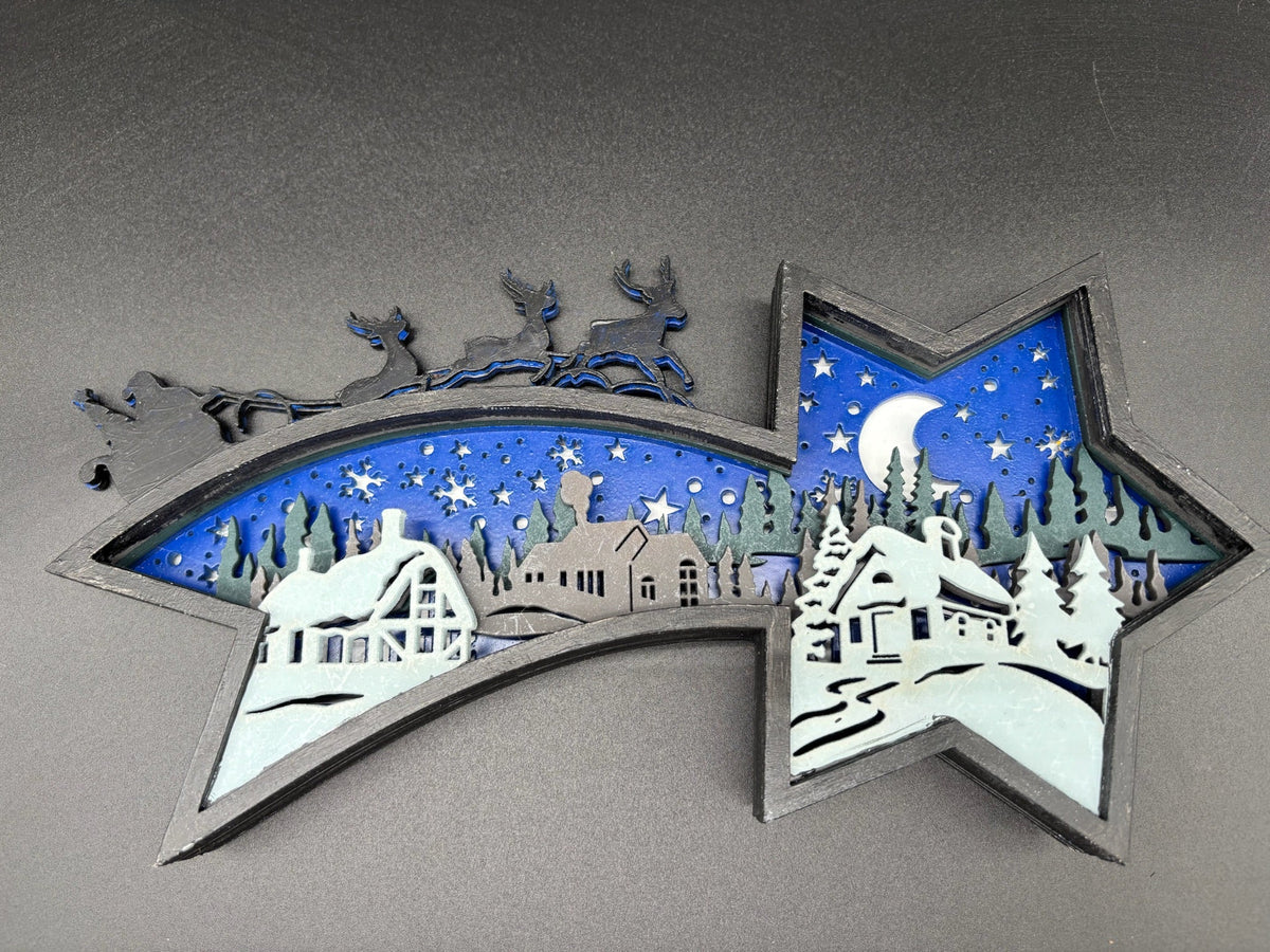 A star-shaped decorative wall art piece featuring a layered winter village scene with blue sky, white snow, gray buildings, and a crescent moon, mounted on a gray textured wall.