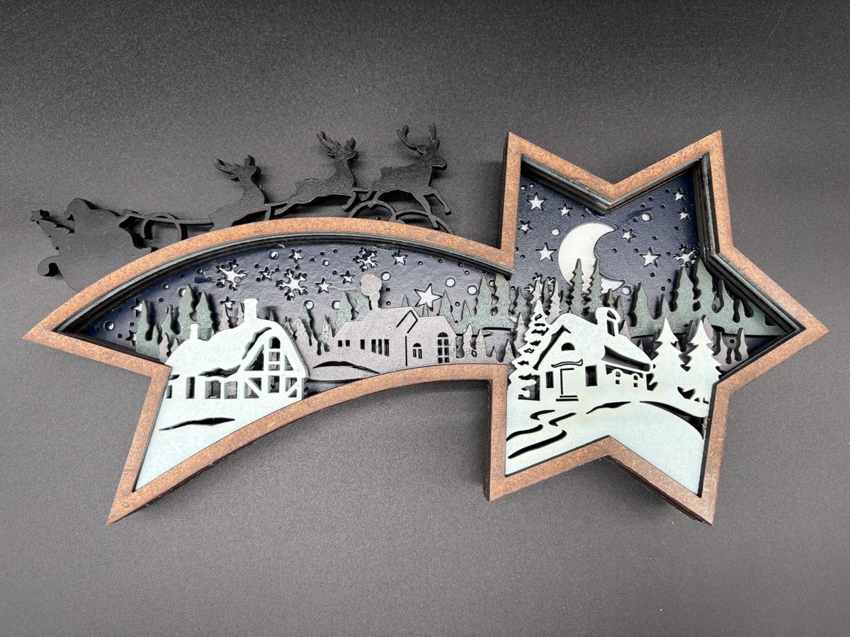 A star-shaped layered paper-cut wall art piece featuring a winter village scene with snow-covered houses, trees, stars, and a crescent moon, mounted against a dark gray wall.