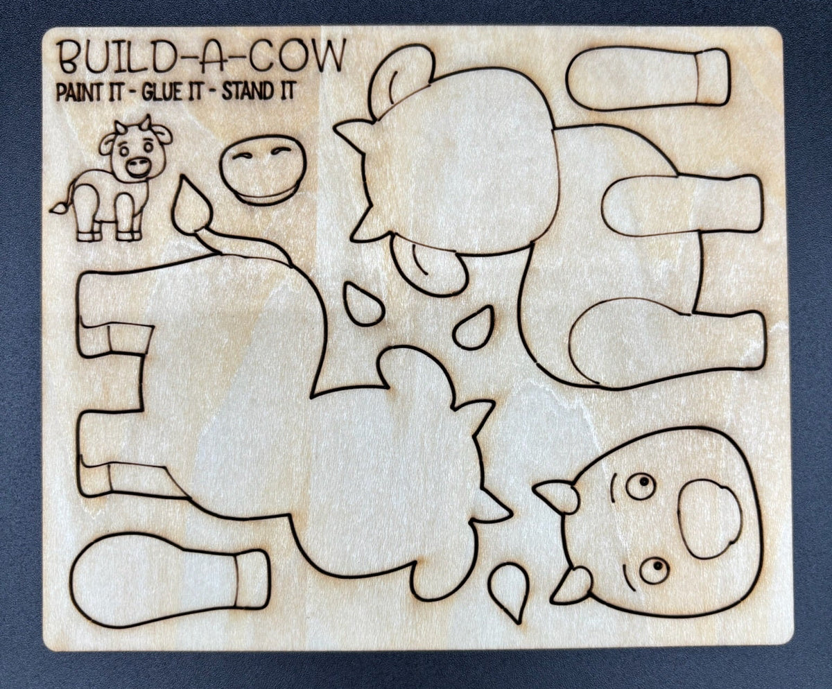 A wooden craft kit labeled 'BUILD-A-COW' showing laser-cut outlines of cow body pieces and facial features on a dark background.